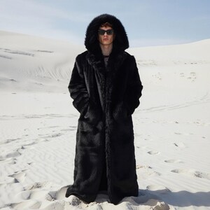 May include: A long, black faux fur coat with a large hood. The coat is worn by a person wearing sunglasses, standing in a desert landscape. The coat has a classic, full-length design.