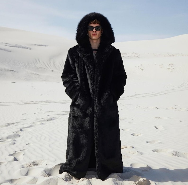May include: A long, black faux fur coat with a large hood. The coat is worn by a person wearing sunglasses, standing in a desert landscape. The coat has a classic, full-length design.