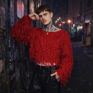 May include: A bright red, fringed sweater with a cropped hem. The sweater has long sleeves and is embellished with sequins. The model is wearing black leather pants and a silver necklace with a cross pendant. The background is a city street.