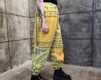 May include: Yellow fleece sweatpants featuring a black bandana-style paisley print pattern. The pants have an elastic waistband with a drawstring, elastic cuffs at the ankles, and a relaxed fit, paired with black platform boots and a black t-shirt.