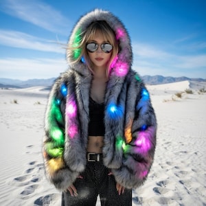 Puede incluir: Chaqueta con capucha de piel sintética gris con luces LED de colores integradas. La chaqueta presenta un cinturón con hebilla plateada y se usa en un paisaje desértico. Las luces son en tonos rosa, verde, azul y amarillo.