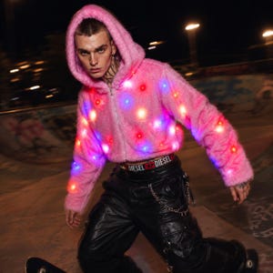 May include: A bright pink fuzzy hooded zip-up jacket featuring integrated LED lights that glow in red and blue. The jacket has long sleeves, a hood, and a front zipper closure. The fabric has a textured, faux-fur appearance throughout the garment.