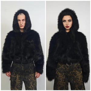 May include: A black faux fur hooded jacket with a zip-up front. The jacket has long sleeves and a cropped length. The model is wearing patterned pants. The jacket is a statement piece.