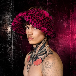 Può includere: Un cappello a secchiello realizzato in morbida pelliccia sintetica con un vivace motivo leopardato rosa acceso e nero. Il cappello presenta una tesa larga inclinata verso il basso e una consistenza soffice che copre l'intera superficie esterna.