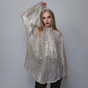 Puede incluir: Una camisa plateada con lentejuelas, mangas largas y cuello camisero. La tela brillante crea un efecto luminoso. La camisa tiene un corte holgado y puños abotonados. Una prenda elegante para la noche.
