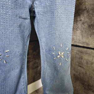 May include: Light blue denim jeans with a pattern of small holes and scattered rhinestones. The jeans feature rhinestone floral designs on the lower legs. The fabric has a faded appearance.