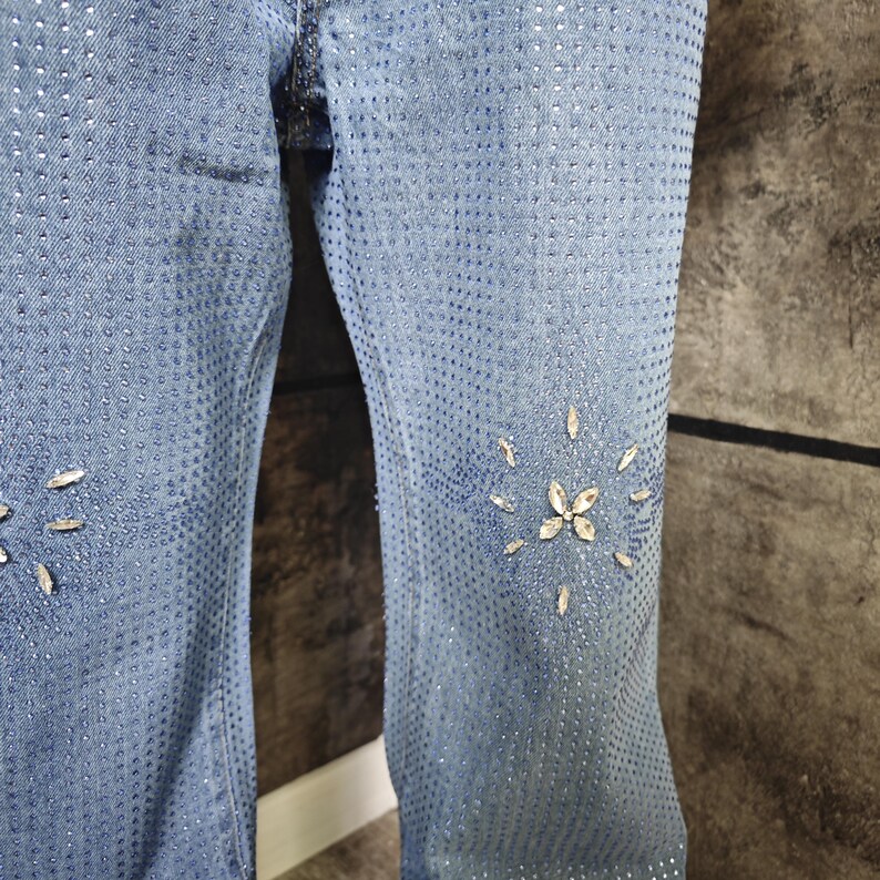 May include: Light blue denim jeans with a pattern of small holes and scattered rhinestones. The jeans feature rhinestone floral designs on the lower legs. The fabric has a faded appearance.
