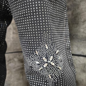 May include: Close-up of a black garment sleeve densely covered in small, silver-colored embellishments. A floral design made of larger, clear crystals adorns the lower sleeve, adding a touch of sparkle. The fabric appears to be a dark denim or similar material.