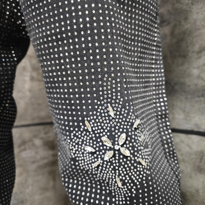 May include: Close-up of a black garment sleeve densely covered in small, silver-colored embellishments. A floral design made of larger, clear crystals adorns the lower sleeve, adding a touch of sparkle. The fabric appears to be a dark denim or similar material.