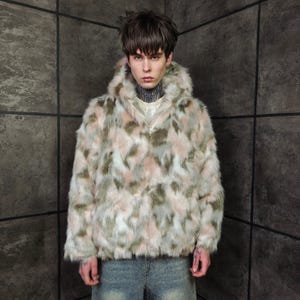 Könnte beinhalten: Kapuzenjacke aus Kunstfell mit einem Camouflagemuster aus Rosa, Grün und Weiß. Die Jacke hat einen Reißverschluss und ein weiches, strukturiertes Aussehen. Das Model trägt ein weißes Hemd darunter und blaue Jeans.