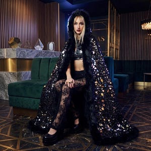 Puede incluir: Una capa negra con capucha con un ribete de piel sintética y un diseño de espejo. La capa se usa sobre un top negro y pantalones de encaje. La persona está sentada en un sofá de terciopelo verde.