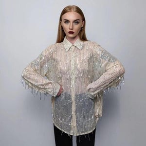 Puede incluir: Una camisa transparente color crema con botones y mangas largas. La camisa está adornada con flecos y lentejuelas plateadas verticales, creando un efecto brillante. Tiene un cuello y puños clásicos.