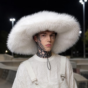 Op de afbeelding: Een oversized witte hoed van imitatiebont met een brede rand en een dunne zwarte kinband. De hoed wordt gecombineerd met een wit shirt met textuur, voorzien van brede schouderbanden en metalen hardware voor een monochrome uitstraling.