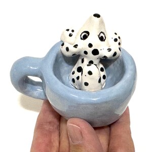 May include: A ceramic figurine of a Dalmatian dog with black spots sits inside a light blue teacup. The dog has black eyes and ears. The teacup has a handle and a smooth, glossy finish. The figurine is a whimsical decorative item.