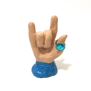 May include: A ceramic hand sculpture in a blue and white speckled design, making the "rock on" hand gesture. A turquoise ring with a large, faceted stone is displayed on the index finger.
