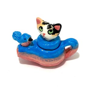 May include: A whimsical ceramic teapot in the shape of a cat with a blue scarf and a bluebird perched on the spout. The teapot is painted in shades of blue, pink, and orange, with a calico cat head as the lid.