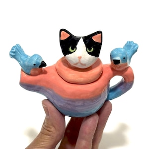 May include: A whimsical ceramic teapot featuring a black and white cat head with green eyes, a pink collar, and two blue bird handles. The teapot is painted with a gradient of pink, orange, and purple hues.