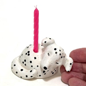 May include: A white ceramic Dalmatian dog candle holder with black spots. A bright pink candle is inserted into the dog's back. The candle holder is a decorative item.