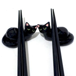 May include: Two black ceramic cat chopstick rests with pink inner ears and green eyes. The chopstick rests are positioned on either side of a pair of black chopsticks. The cats are facing each other, creating a charming and functional dining accessory.