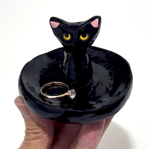 May include: A black ceramic ring dish in the shape of a cat. The cat has yellow eyes and pink ears. A gold ring with a clear stone rests in the dish. The dish is held by a hand.