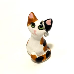 May include: A ceramic calico cat figurine with white, orange, and black patches, green eyes, and pink inner ears. A silver-toned ring with a clear, round faceted stone is placed over one of the cat's ears, serving as a decorative ring holder.