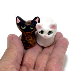 May include: Two ceramic cat figurines. One is a calico cat with black, orange, and brown markings. The other is a white cat. Both have green eyes and pink accents on their ears and noses. The figurines are small and appear to be handmade.