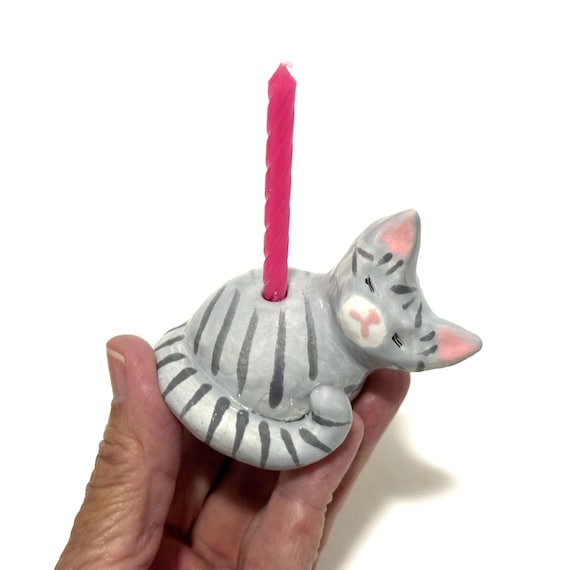 A hand-sculpted ceramic gray tabby cat that doubles as a birthday cake topper AND a candle holder — genuinely charming dual-function craft.