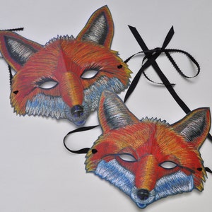 Puede incluir: Dos máscaras de zorro hechas de papel con lazos de cinta negra. Las máscaras son de color naranja, rojo y blanco con ojos negros y un hocico blanco.