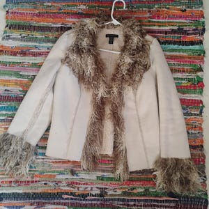 May include: Cream-colored jacket with faux fur trim on the collar and cuffs. The jacket is hanging on a white hanger against a colorful woven rug background. The jacket has a tailored fit and a textured appearance.