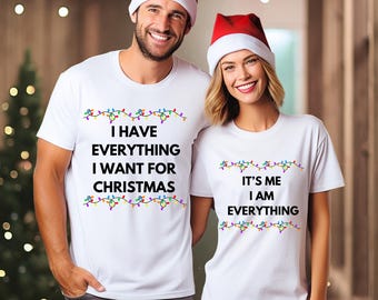 May include: Two people wearing matching white t-shirts with Christmas-themed text and colorful string lights. One shirt reads "I HAVE EVERYTHING I WANT FOR CHRISTMAS," and the other says "IT'S ME I AM EVERYTHING." Both are wearing Santa hats.