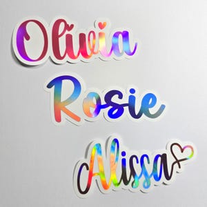 May include: Three holographic name stickers: "Olivia," "Rosie," and "Alissa." The names are in a cursive font with a rainbow sheen, outlined in white. A small heart appears above the "i" in "Olivia" and next to the "a" in "Alissa."
