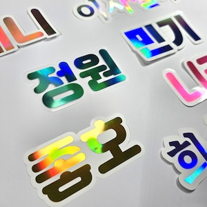 May include: A collection of holographic stickers with Korean text. The stickers feature various colors, including green, blue, pink, and gold. The stickers are on a white background.
