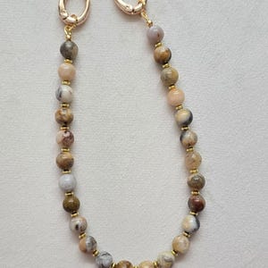May include: A beaded chain featuring round, multi-colored earth-toned stones in shades of brown, beige, gray, and cream. The beads are separated by small gold-colored metal spacers and the chain ends with two gold-colored metal lobster clasps.