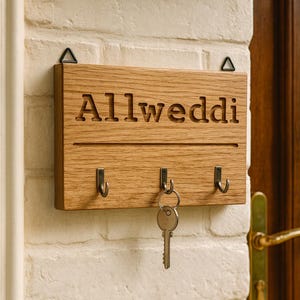 May include: Wooden key holder with the word "Allweddi" carved into the wood. The key holder has three hooks and a key hanging from the center hook. The key holder is mounted on a white brick wall.