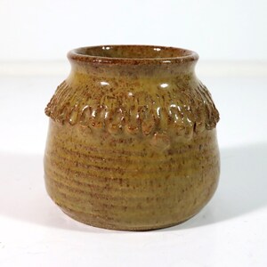 May include: A small, handcrafted ceramic vase with a textured, earthy brown glaze. The vase features a decorative band of raised, rounded details near the top, adding visual interest. The overall shape is rounded with a slightly flared rim.