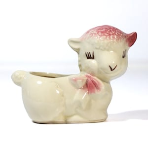May include: A white ceramic planter shaped like a lamb with pink ears and a pink bow. The lamb has closed eyes and a sweet expression.