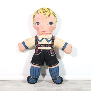 May include: A vintage cloth doll with a painted face, blue eyes, and blonde hair. The doll is dressed in a cream-colored shirt with blue trim, black shorts, and blue socks. The shorts and suspenders are decorated with red floral accents.
