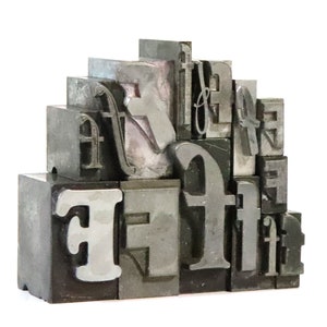 May include: A collection of vintage metal letterpress printing blocks, stacked in a pyramid shape. The blocks are made of lead and feature the letters "F" and "f" in various styles.