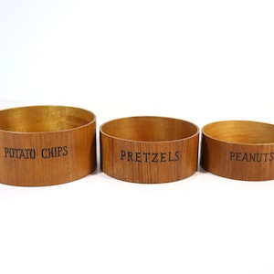May include: Three wooden bowls with labels for potato chips, pretzels, and peanuts.