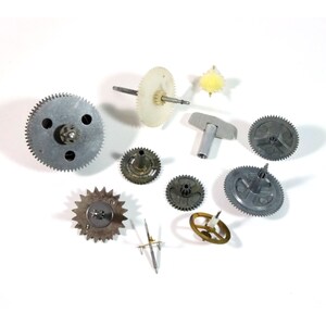 May include: A collection of various metal clock gears and components, including circular gears with teeth, a star-shaped gear, and a small yellow gear. The gears are in shades of silver, gold, and off-white, scattered on a white surface.