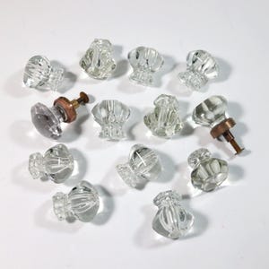 May include: A collection of clear glass cabinet knobs with various designs. Some knobs have a faceted, rounded shape, while others have a more ornate, textured appearance. Two knobs have brass hardware attached. The knobs are arranged on a white surface.