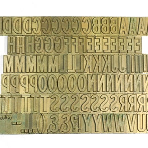 May include: A collection of brass letterpress printing blocks, including uppercase letters, numbers, and punctuation marks. The blocks are arranged in rows and columns, with the letters A, B, C, D, E, F, G, H, I, J, K, L, M, N, O, P, Q, R, S, T, U, V, W, X, Y, Z, 0, 1, 2, 3, 4, 5, 6, 7, 8, 9, !, ?, and - visible.