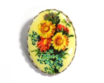 May include: A yellow oval brooch with a floral design. The flowers are yellow, red, and blue.