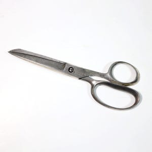 Puede incluir: Un par de tijeras de metal vintage con grandes orificios para los dedos y hojas largas. Las tijeras son de color plateado y muestran signos de desgaste. Están diseñadas para cortar tela o papel. Las hojas miden unos 20 cm de largo.