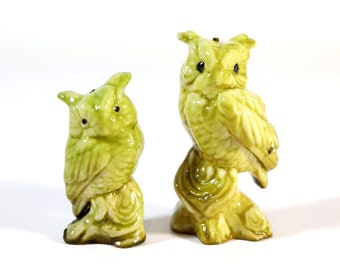 May include: Two ceramic owl figurines, both green with brown eyes. The owls are perched on a branch and have a detailed design.