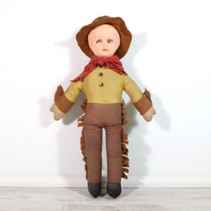 May include: A vintage cowboy doll with a painted face, wearing a brown hat, red scarf, yellow shirt, brown pants with fringe, and brown arm cuffs. The doll has black boots and is standing against a white background.