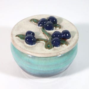 May include: A ceramic jar with a lid, featuring a teal base and a cream-colored lid. The lid is adorned with a raised design of dark blue berries and green leaves. The jar is a decorative piece.