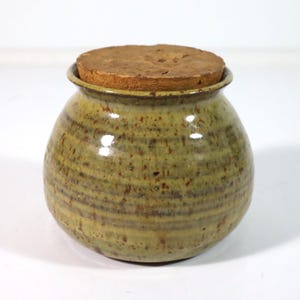 May include: A handmade, round ceramic jar with a cork lid. The jar has a speckled, earthy green and brown glaze. The cork lid is a light brown color. The jar is a decorative storage container.