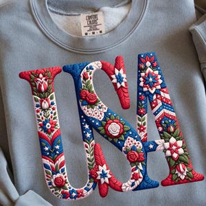 Peut inclure: Un sweat-shirt bleu clair avec le mot "USA" en design floral rouge, blanc et bleu. Les lettres sont ornées de fleurs et de motifs. Le sweat-shirt a un col rond et des manches longues.