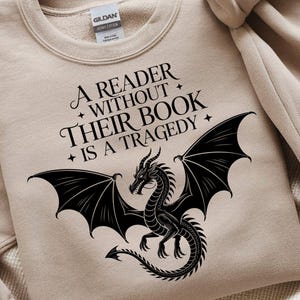 Peut inclure: Un sweat-shirt beige avec le texte "A READER WITHOUT THEIR BOOK IS A TRAGEDY" et un dessin de dragon noir. Le dragon a de grandes ailes et une longue queue. Le sweat-shirt a un col rond et des manches longues.
